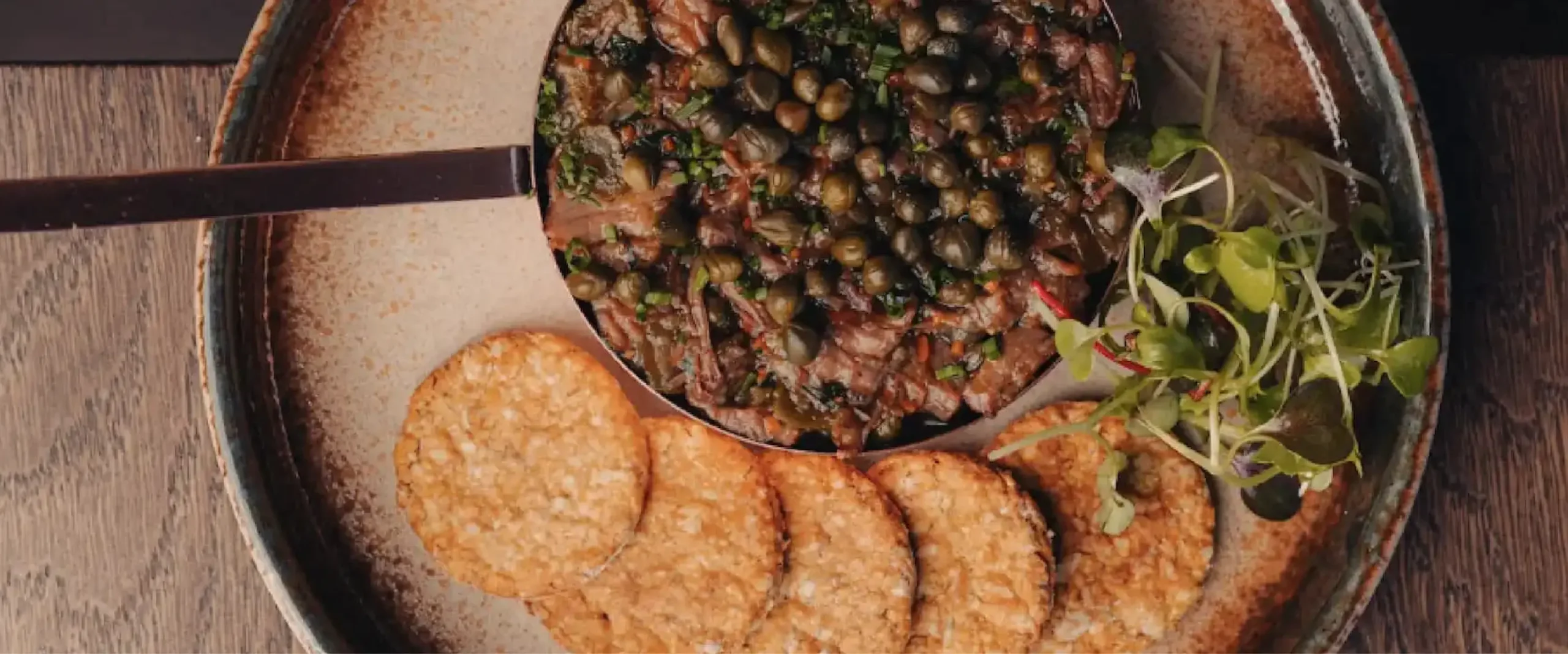 Cast-iron skillet with a clam and meat hash topped with herbs, beside round flatbreads on a wooden table.