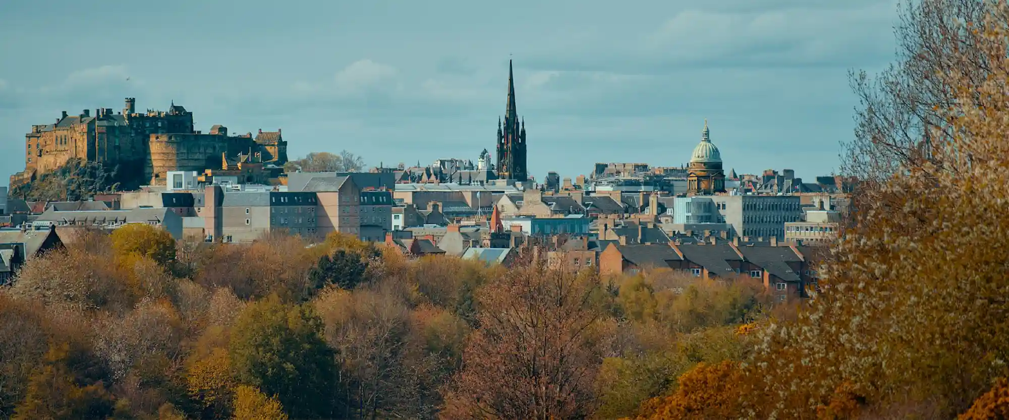 Panoramic cityscape with a castle on the left hill, a Gothic spire in the center, a domed building on the right, and autumn trees in the foreground.