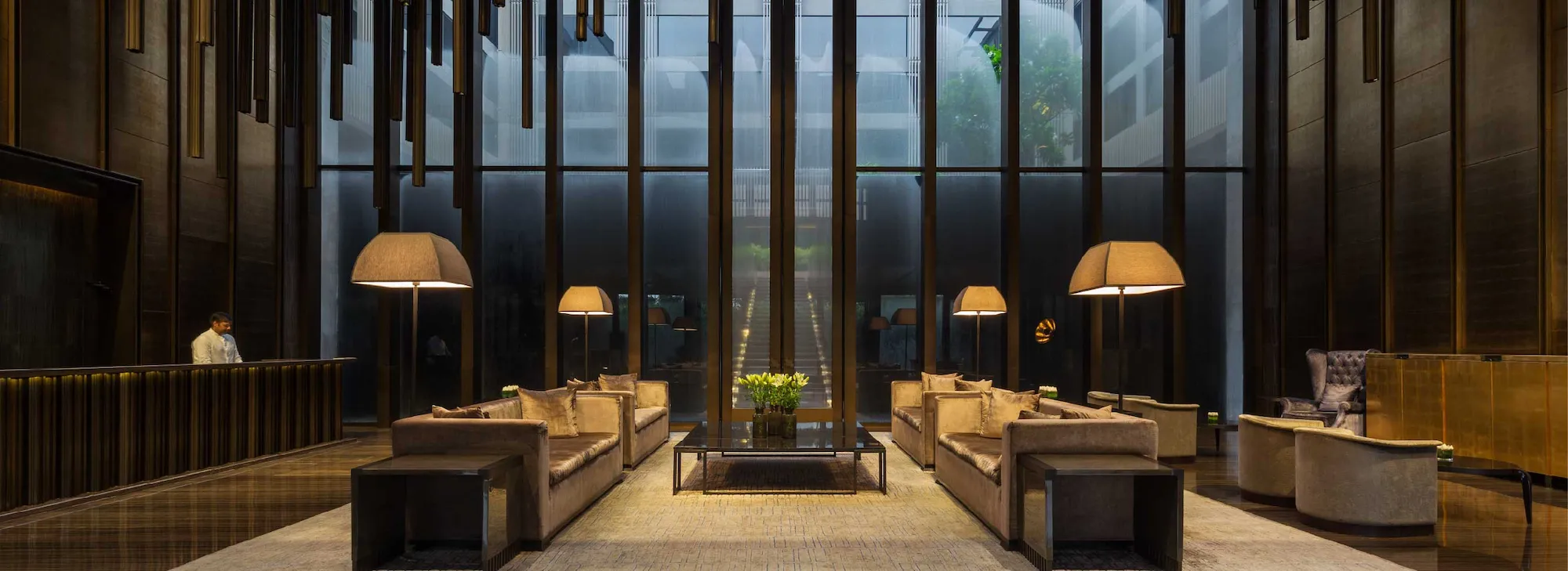 Hotel lobby with a dark wood reception desk on the left and beige sofas arranged around a glass coffee table, tall windows behind.