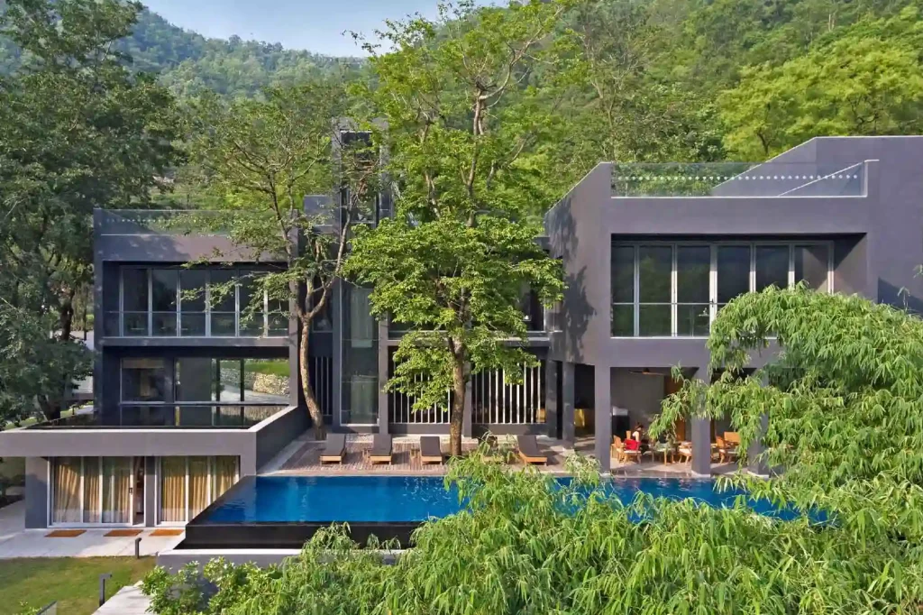 Modern gray villa with large glass windows, pool, and lounge chairs among trees on a hillside backdrop.