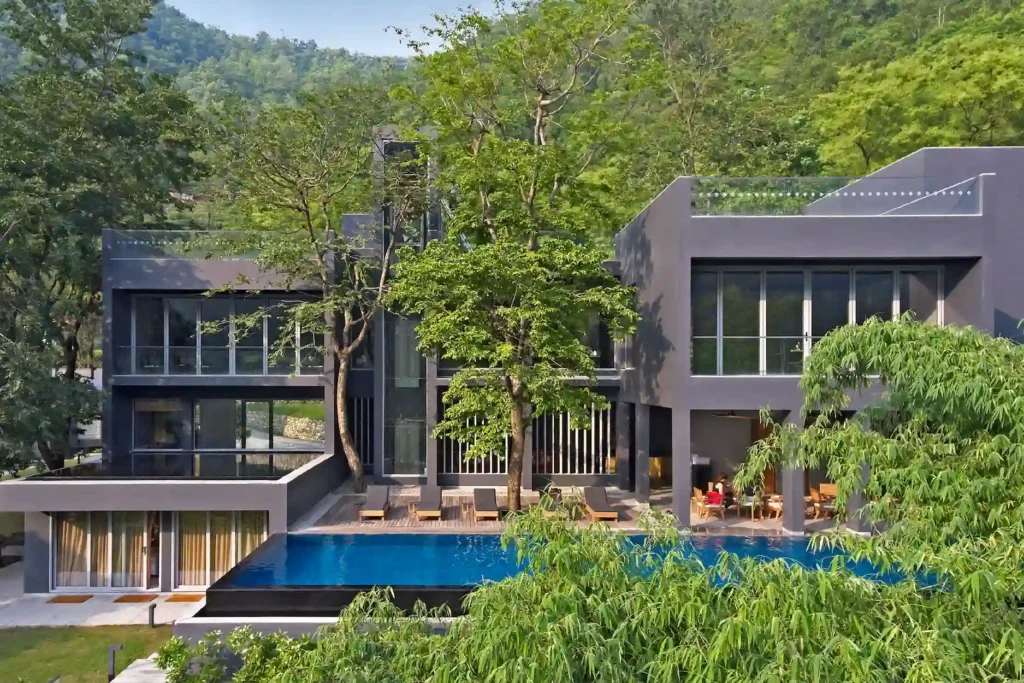 Modern gray villa complex with glass railings, overlooking a rectangular pool and lush trees; hillside in the background.
