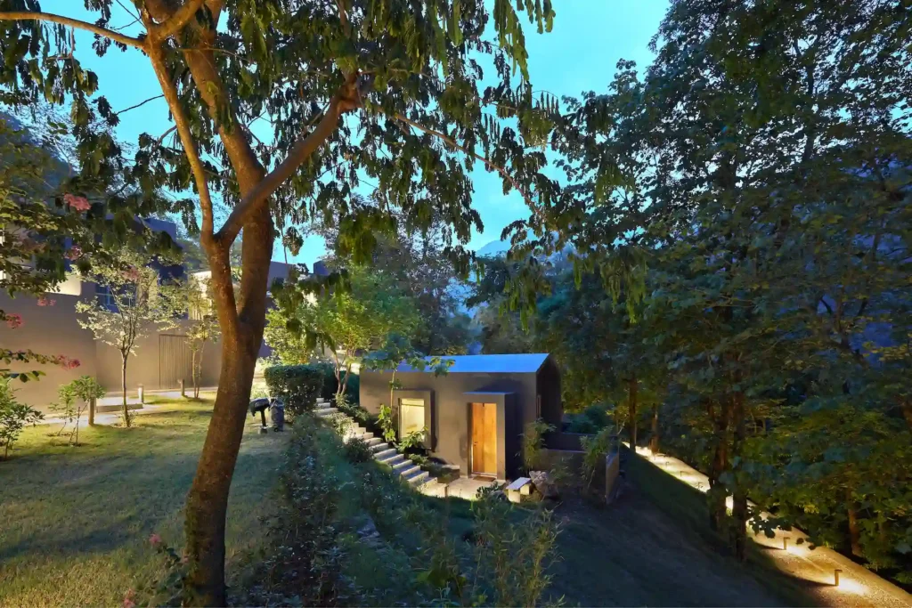 card-one | The Roseate Ganges Rishikesh Twilight view of a modern house with wooden doors, surrounded by trees and a lit pathway in a landscaped garden