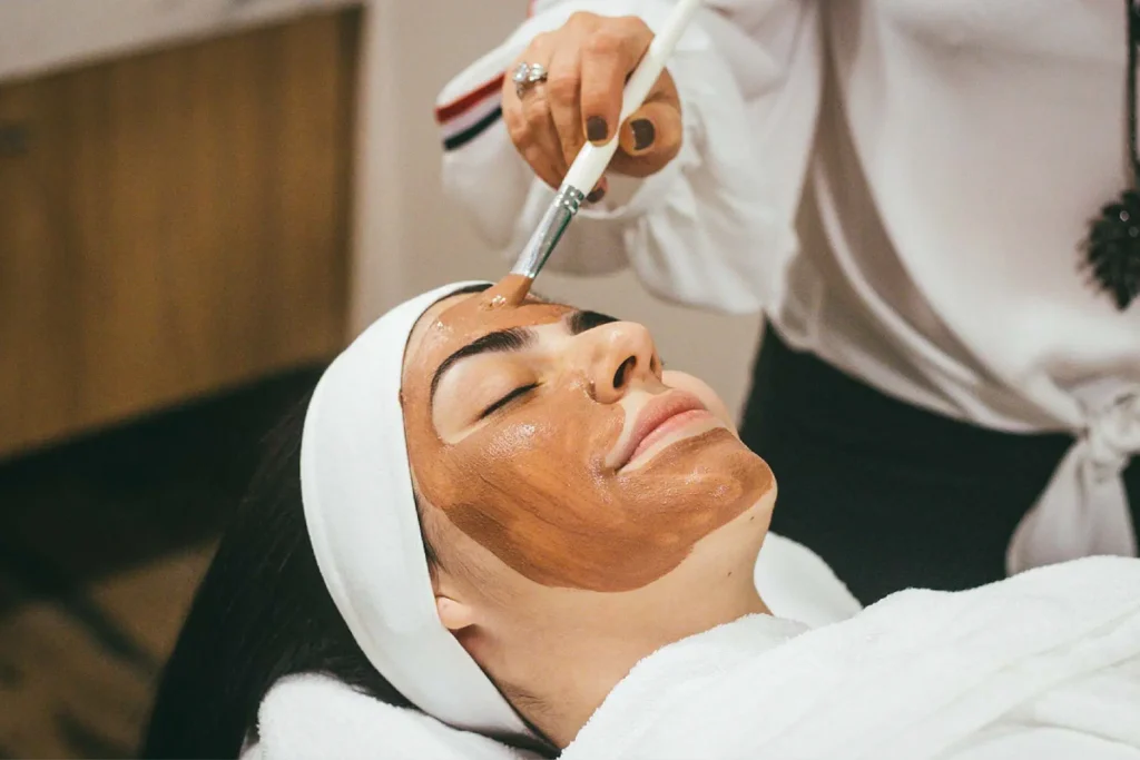 skin-needs-attention | The Roseate Ganges Rishikesh Woman lying with a brown facial mask as a spa professional applies it with a brush.