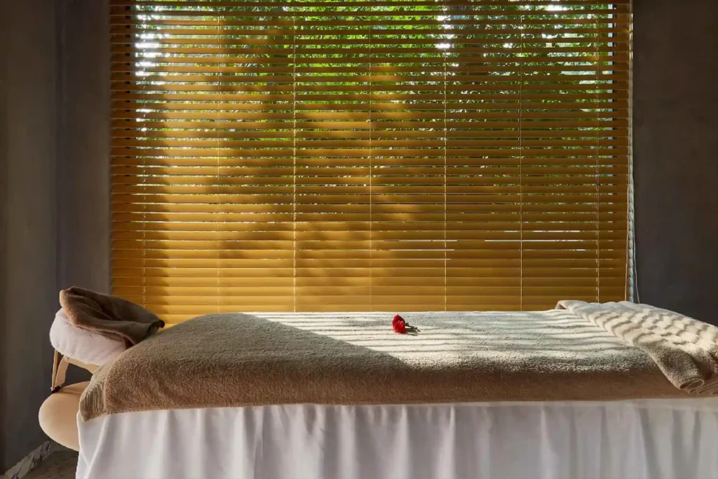 wellbeing-at-aheli | The Roseate Ganges Rishikesh Massage bed with beige blanket and a single red flower, set against sunlit wooden blinds.